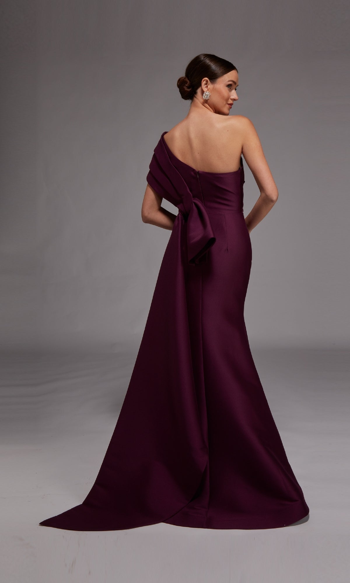 A woman models the Alyce 27838 long formal dress—deep purple, one-shoulder style with a long train and large back bow—standing against a plain gray background, highlighting the gown’s elegance.